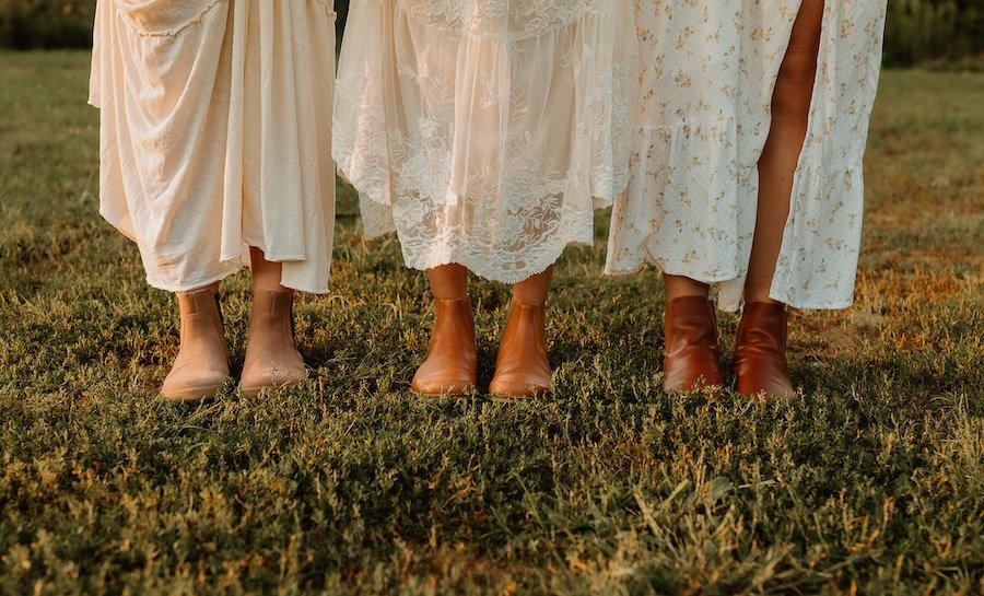 A line of 3 light brown barefoot chelsea boots being worn by 3 women in white and cream colored dresses. All 3 boots feature a wide toe box. Left to right are Be Lenka Entice, Lang S Luna, and Rosa by Anya
