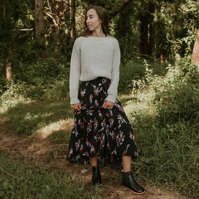 A full body view of a woman standing on a nature path wearing a cream sweater and a long black floral skirt. She is holding the hem up slightly to show off a pair of black leather wide toe box chelsea boots: The Lila by Anya's Shop.