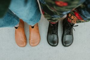 A top down view of two pairs of feet standing on concrete standing side by side wearing denim and Lang.S barefoot boots - the Luna chelsea in caramel on the left and the Kombat in black leather on the right to illustrate the wide anatomical shape of the boots