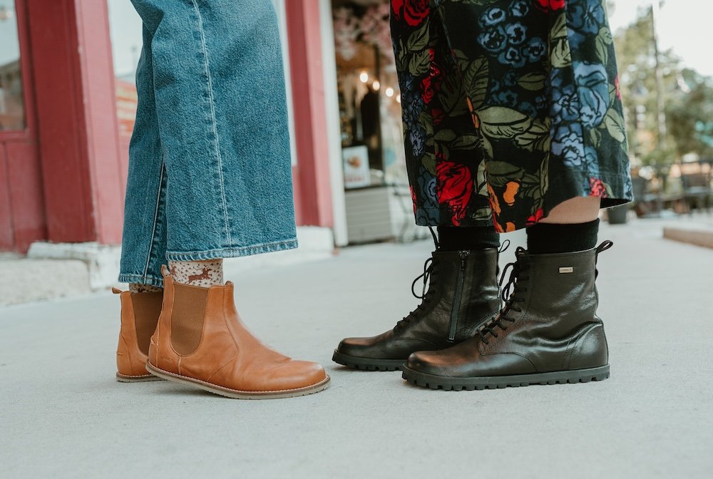 A ground level view of two pairs of feet standing on concrete facing each other wearing denim and Lang.S barefoot boots - the Luna chelsea in caramel on the left and the Kombat in black leather on the right