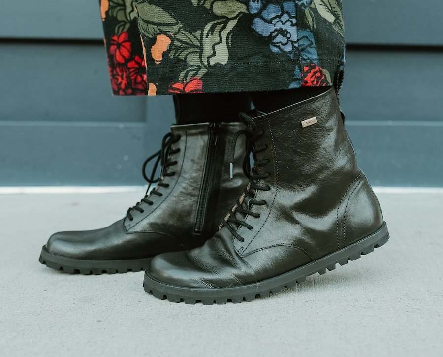 A close up side view of a pair of feet standing on concrete wearing black leather Lang.S barefoot Kombat boots