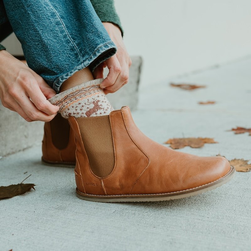 A close up side view of a person's feet resting on concrete with fall leaves around, and hands reach down to adjust the socks. The feet are wearing Lang.S Luna caramel leather barefoot chelsea boots