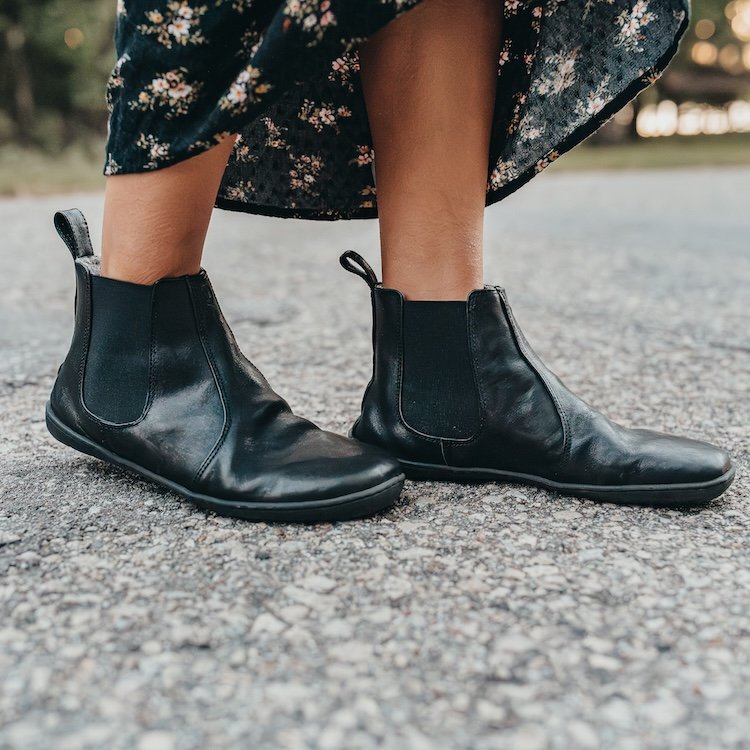 A zoomed in view of a woman walking on a stone pathway wearing a long black floral skirt paired with Mukishoes black leather barefoot chelsea boots.