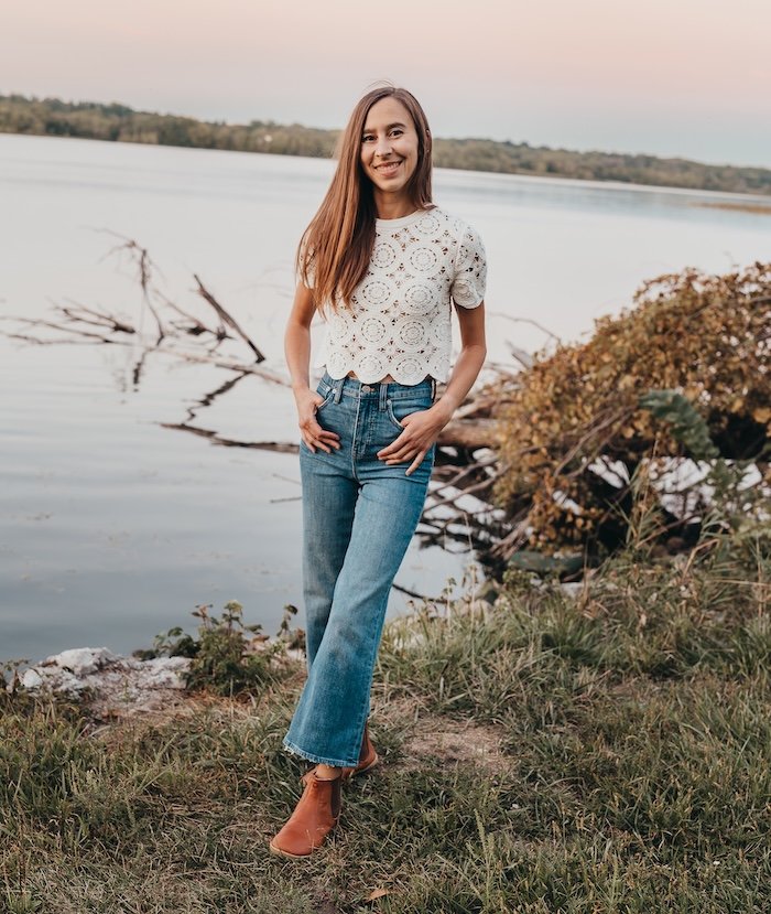 A full body view of a woman wearing Mukishoes brown leather barefoot chelsea boots with a crochet top and mid tone denim. She has her thumbs hooked in her pockets and is smiling at the camera standing near a lake.