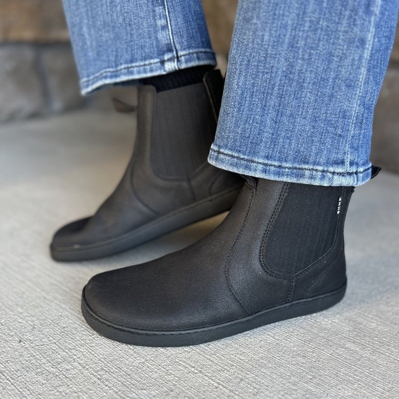 A pair of vegan chelsea boots in black from Bohempia being worn on cement. They are shown from a side angle with the back foot flexed up to demonstrate flexibility of the zero drop outsole