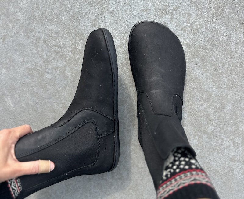 A top down view of a pair of feet resting on pavement wearing colorful winer socks and Groundies Barefoot+ wide toe box black nubuck chelsea boots