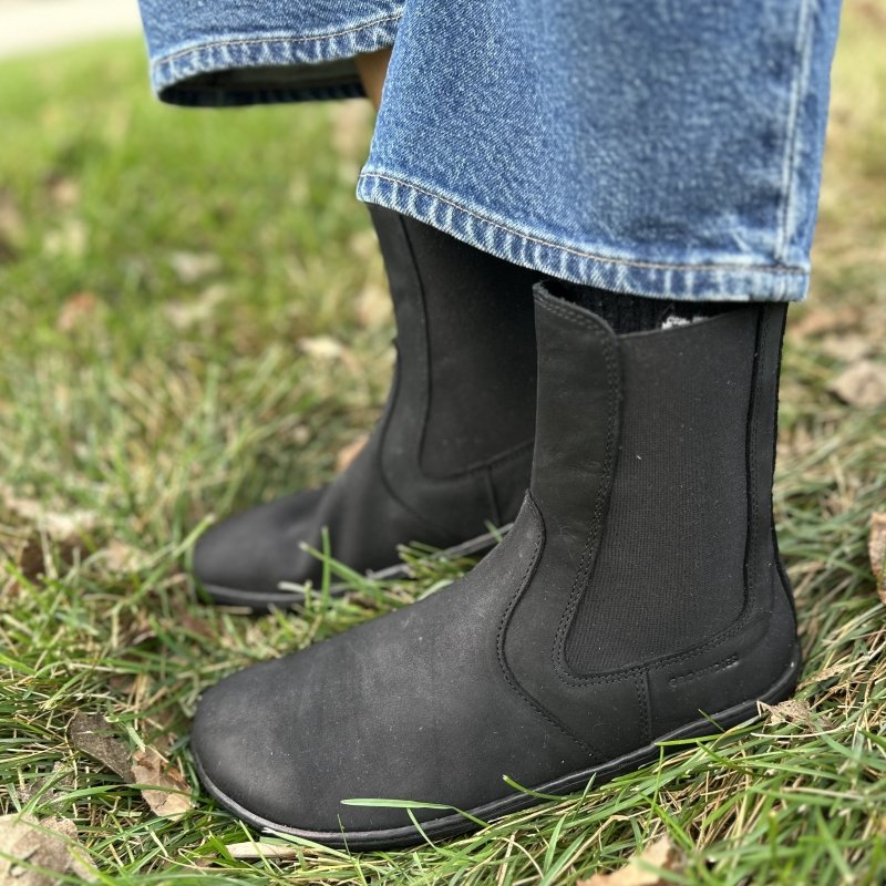 A side view of a person standing on grass with wide leg jeans resting above their boots, the Groundies Camden in Barefoot+ width