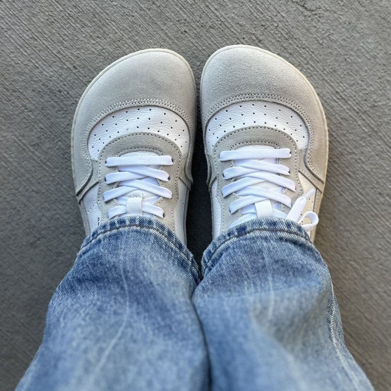 Top down view of a pair of feet in Groundies Barefoot Plus Nova sneakers with an extra wide toe box. They are gray and white and shown with medium wash denim.