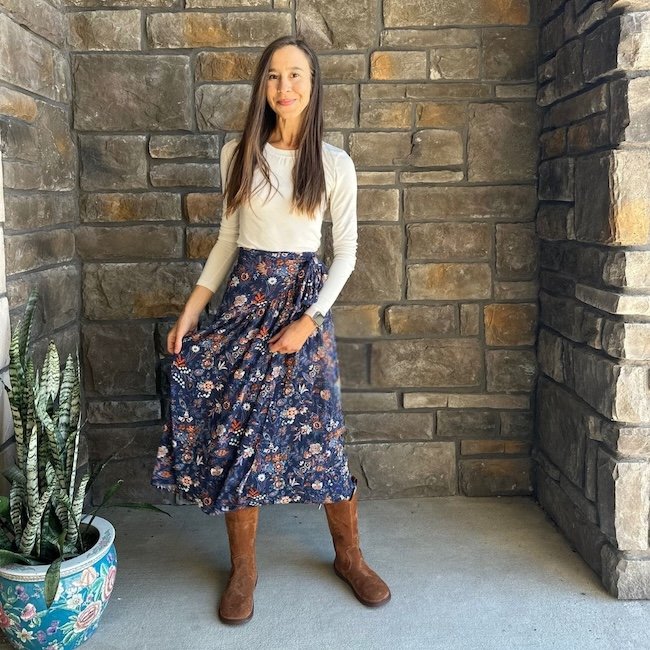 A full body view of a woman in a long sleeve cream shirt, navy floral flowy skirt, and Groundies Tuscon barefoot cowboy style boots with a wide toe box.