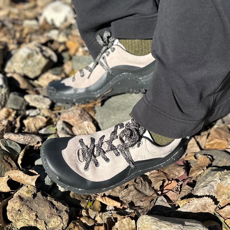 A pair of Lono Wild hiking shoes on rocky ground. They are black and gray with speckled laces and a wide toe box
