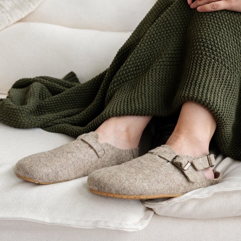 A person's legs covered by a cozy blanket, wearing a pair of Mukishoes Soca Lana wool slipper clogs with a comfortable wide toe box