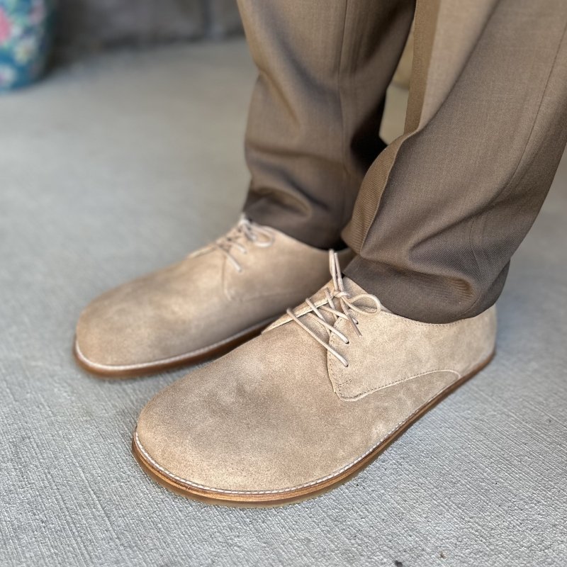 A front angled view of a man's legs from the shin down wearing brown dress pants paired with tan suede barefoot dress shoes from Origo Shoes. The wide toe box Men's Derby has visible stitching around the sole for a classic look