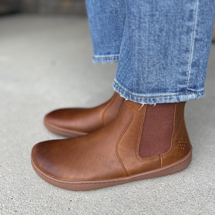 A side view of Vivobarefoot brown leather Sensus chelsea boots with a wide toe box and flat outsole. A pair of jeans are resting at the top of the boots