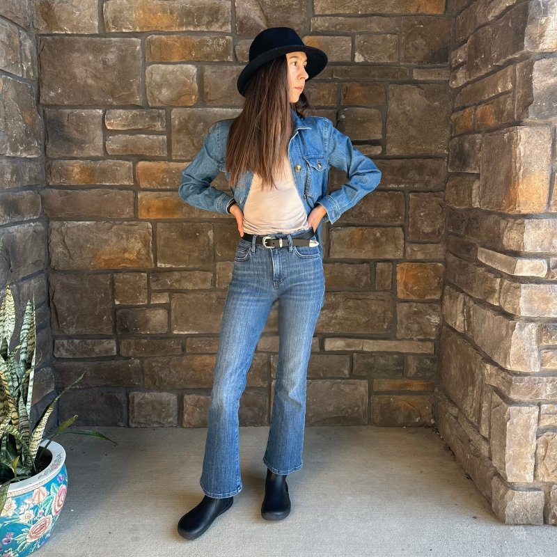 A full body view of a woman in jeans, a white shirt, and a denim shirt. She has accessorized with a black hat, black leather belt, and a pair of black leather Vivobarefoot Sensus chelsea boots. She is standing with her hands on her hips look off to the right.