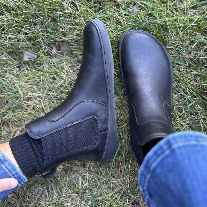 A pair of feet in balck leather Vivobarefoot Sensus Chelsea boots. The right foot is sitting upright to show off the wide toe box and the left foot is laying to the side to show the side profile and zero drop outsole