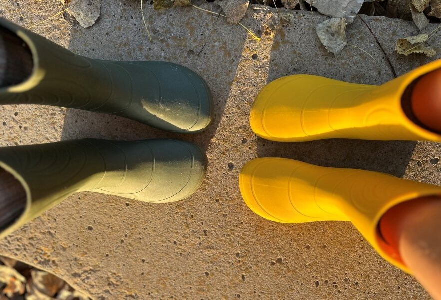Two pairs of feet, one adult and one kid, standing toe to toe while both wearing Wildling Ranidae wide toe box rain boots with a zero drop sole. Olive green and yellow colors are shown