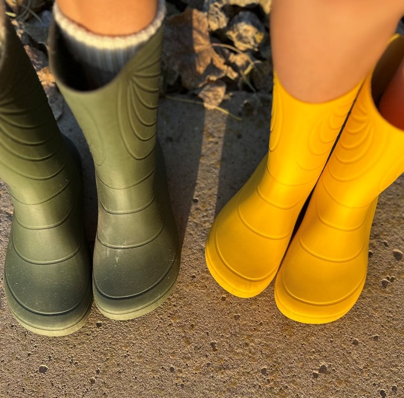 Two pairs of feet in Wildling Ranidae rain boots standing outside. The child's pair on the right is vibrant yellow barefoot wide toe box, and the adult pair features a similar wide toe box in hunter green