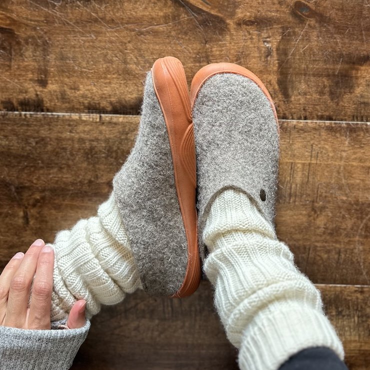 Top down and side view of a pair of Wildling Shoes Slinka Sten wool barefoot clog style slippers with a wide toe box and rubber outsole. The person is also wearing chunky wool leg warmers with them.