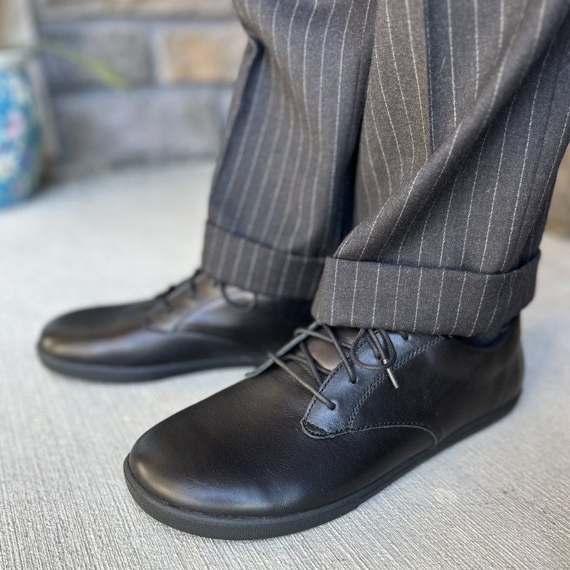 A side view of Xero Glenn, a barefoot dress shoe in black leather worn with gray pin striped pants.