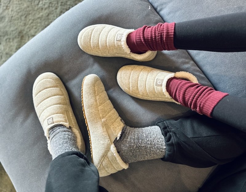 A top down close up view of two people's legs relaxing on a couch. They are both wearing Xero Shoes Pagosa barefoot slippers with a wide toe box and collapsible heel