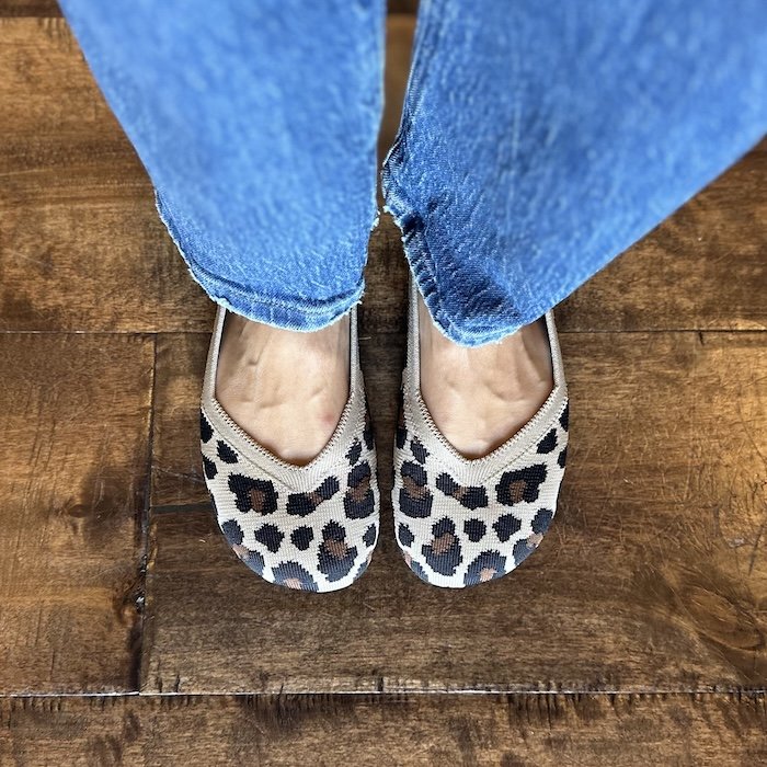 A top down view of a pair of feet standing on wood floor wearing jeans and wide toe box Xero Shoes barefoot ballet flats in leopard print, knit material