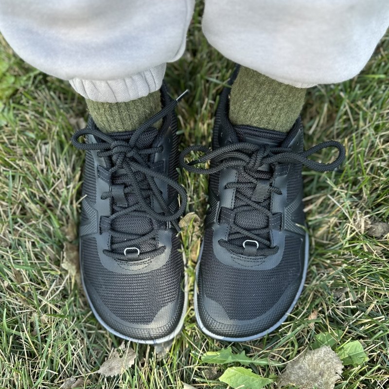 Top down view of Xero Shoes Scrambler Low EV being worn on grassy ground. The wide toe box hiking shoes are paired with cream colored easy pants and green wool socks