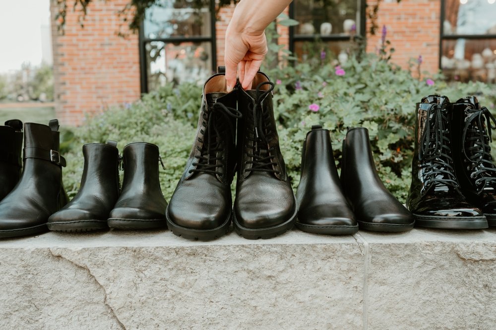 A line up of black barefoot boots resting on a ledge. There is a hand reaching down grabbing the middle pair