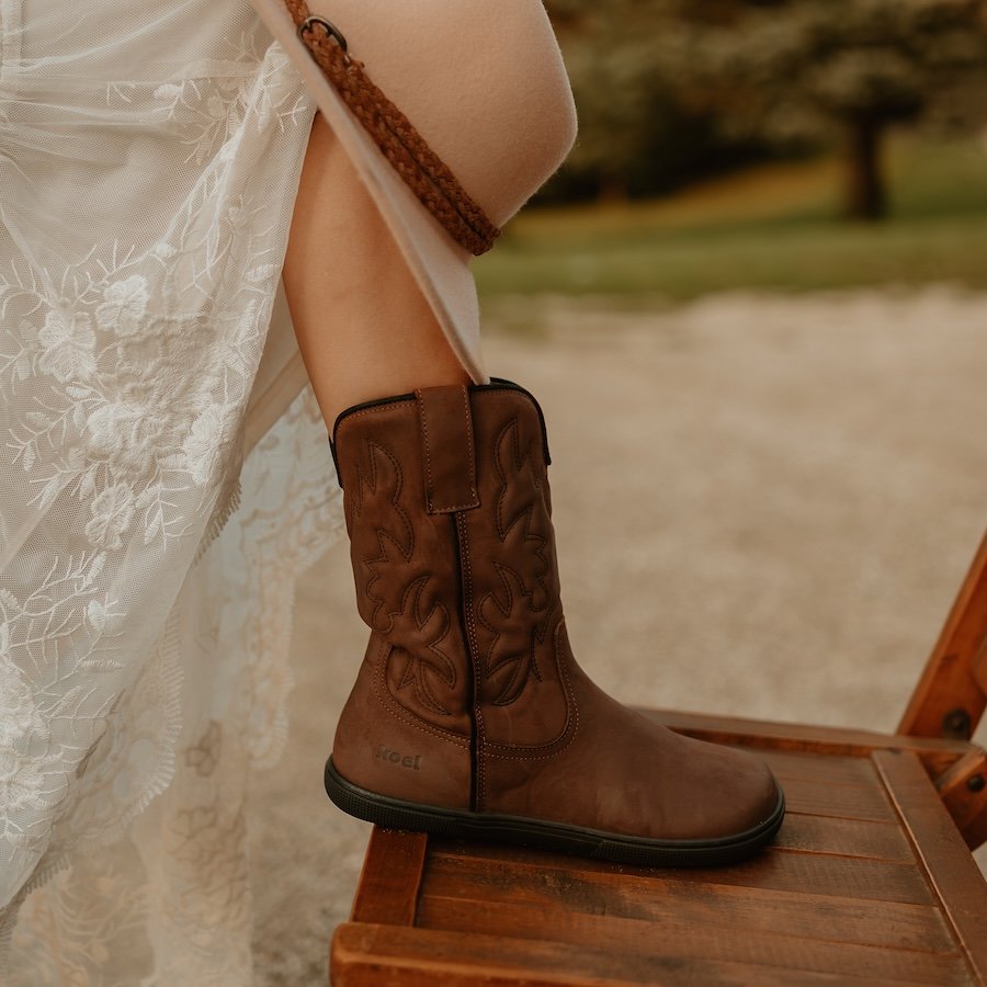 One Koel Fiona Boot resting on an antique wooden chair outside. The person wearing the boot has on a flowy lace dress and is holding a suede hat on her knee. There is a farm blurred in the background. The boot is a leather cowboy style barefoot boot from Koel called Fiona