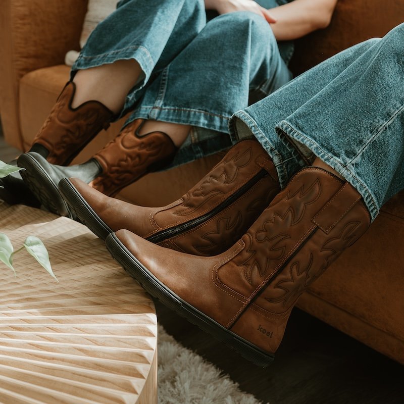 A close up of two women from the knees down sitting on a couch with their feet resting on a coffee table. They are both wearing Koel Fiona barefoot cowboy style boots with a wide toe box in two different colorways - all brown, and black brown