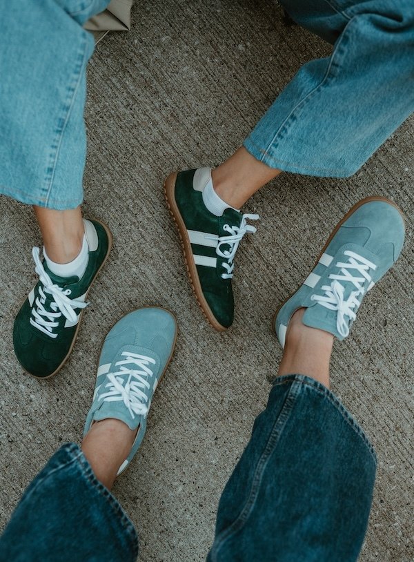 Koel Ila Suede being worn by two people in different colorways. Their feet are casually splayed out on the ground. Both shoes feature a wide toe box and a thin flexible outsole.