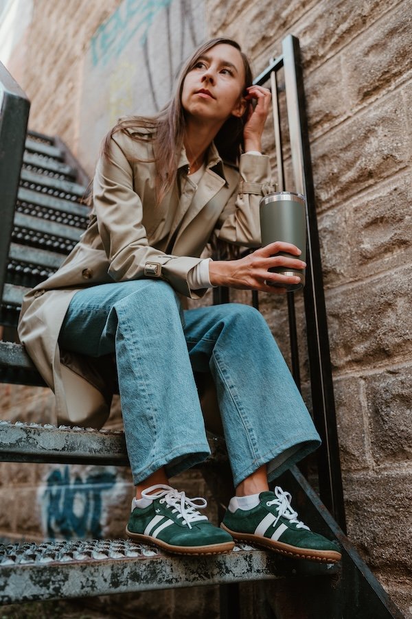 A woman standing on a metal staircase outside a building in jeans and a trench coat. She is staring off into the distance and holding a mug of coffee. On her feet are green Koel Ila Suede sneakers, a barefoot Adidas Sambas alternative