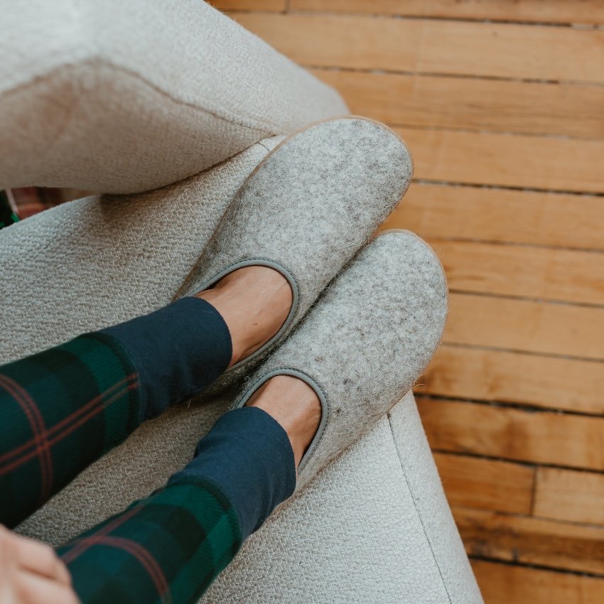 A close up of a pair of feet in Mukishoes Pantufa slippers, a wide toe box barefoot slipper resting on a cozy couch with a wood floor in the background