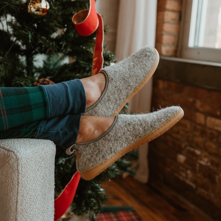 A pair of Mukishoes Pantufa wool cozy barefoot slippers with a wide toe box worn by a person with crossed feet resting on the arm of a couch with a Christmas tree in the background