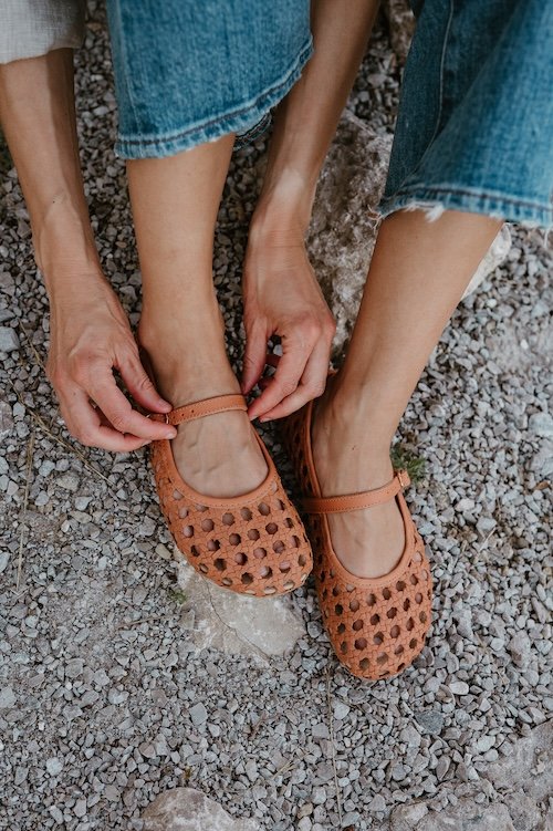 A close up view of a pair of legs and feet resting on gravel outdoors and a pair of hands adjusting the strap on a pair of toffee Origo Shoes x Anya Woven barefoot Mary Janes