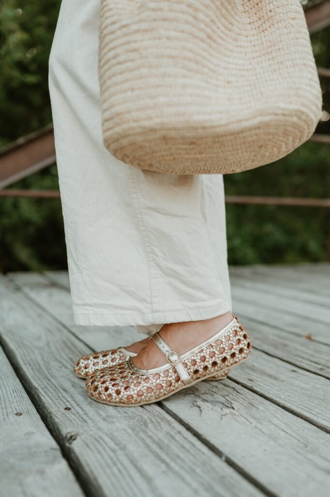 A close up view of a pair of legs and feet standing on a boardwalk outdoors wearing white denim and gold Origo Shoes x Anya Woven barefoot Mary Janes