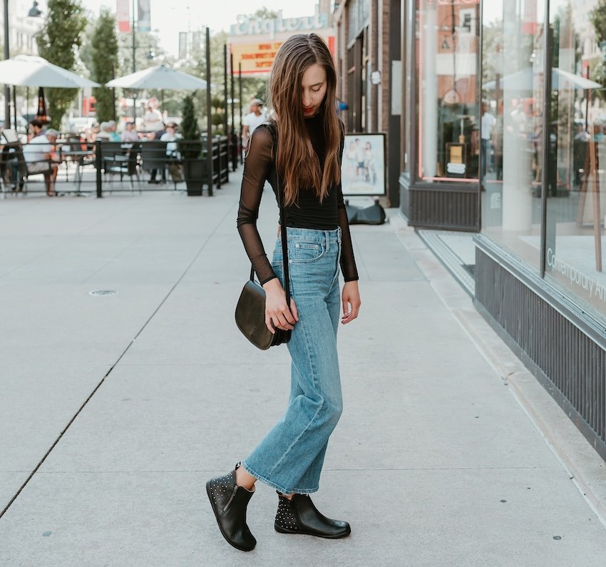 A full body view of a woman in a black mesh long sleeve top, wide leg jeans, and studded Zaqq Qlare barefoot ankle boots with a wide toe box and flexible outsole