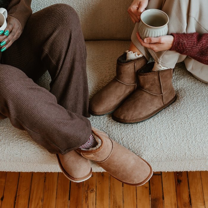A close up cropped image of two people on a cozy sofa wearing Zeazoo Dingo mini sheepskin slippers for adults. They are both dressed in loungewear and holding mugs