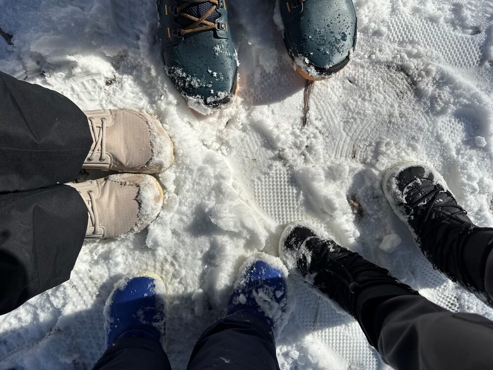 A picture of a family of 4 from the top down, close up on just their boots in the snow. Be Lenka, Vivobarefoot, and Lems barefoot winter boots are all shown