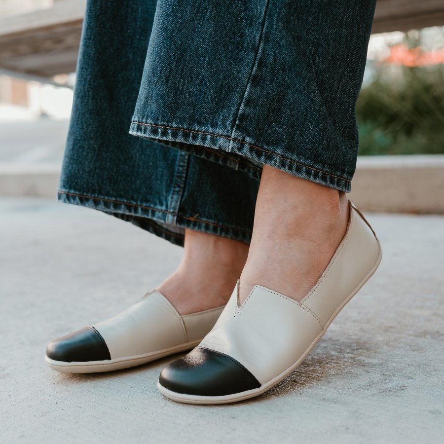 A pair of feet resting on the ground wearing Be Lenka Rosaluna wide toe box flats with a cap toe look. The foot closest to the camera has the heel lifted and a pair of dark wash wide leg jeans are hanging at the ankle.
