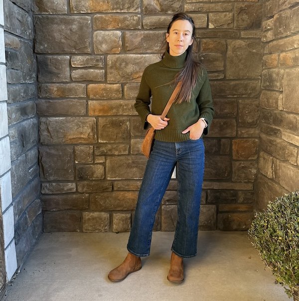 A picture of a woman wearing By Anya Faye wide toe box boots and wide leg jeans and a sweater looking at the camera.