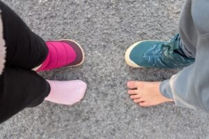 Top down view of 2 kids facing each other. each are wearing 1 snow boot on a foot, and the other foot is bare or socked to show the anatomical toe box of the barefoot Be Lenka and Vivobarefoot snow boots