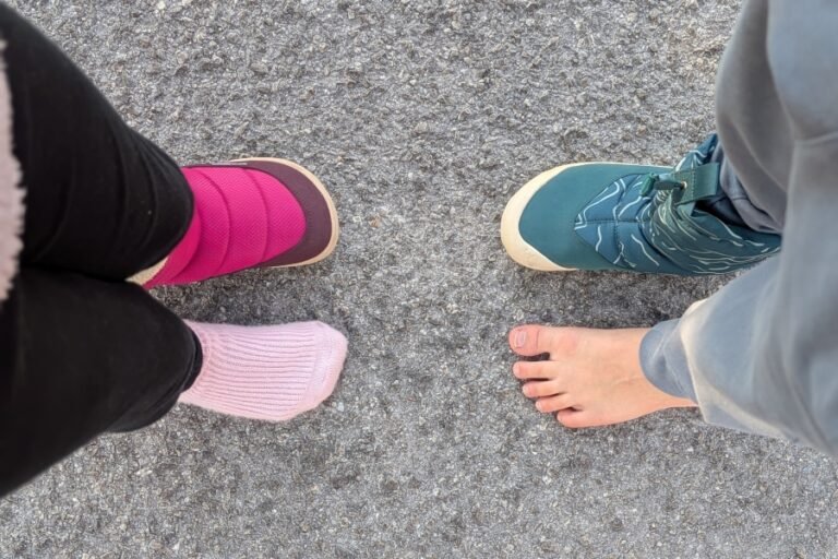 Top down view of 2 kids facing each other. each are wearing 1 snow boot on a foot, and the other foot is bare or socked to show the anatomical toe box of the barefoot Be Lenka and Vivobarefoot snow boots