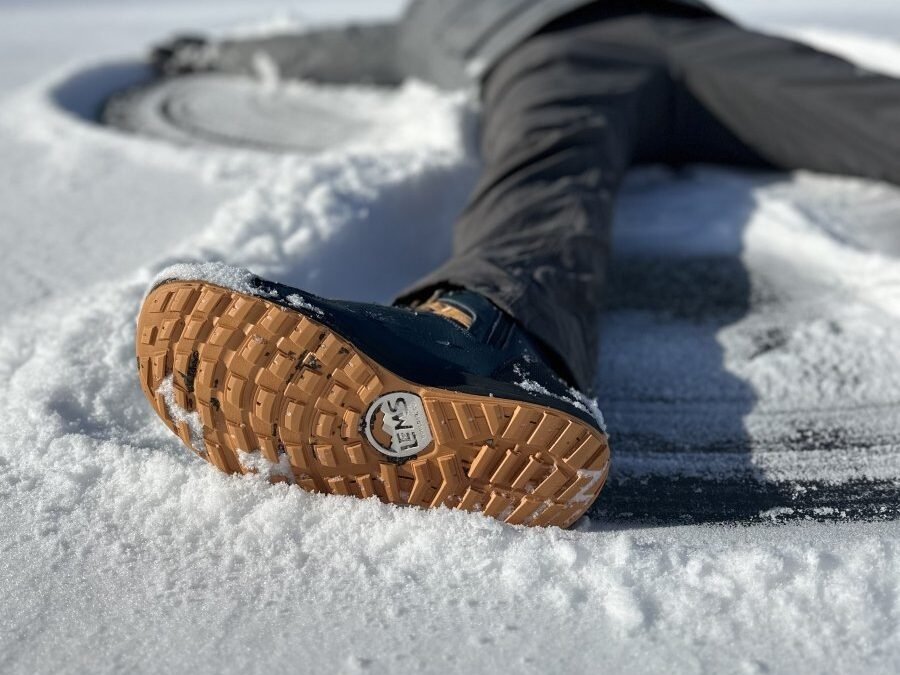 A person laying on the snowy ground making a snow angel. The camera has focused on the outsole of a pair of Lems barefoot winter boots