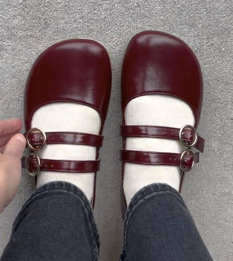 A top down view of a pair of feet standing on pavement wearing burgundy Origo Shoes two strap mary janes with a wide toe box