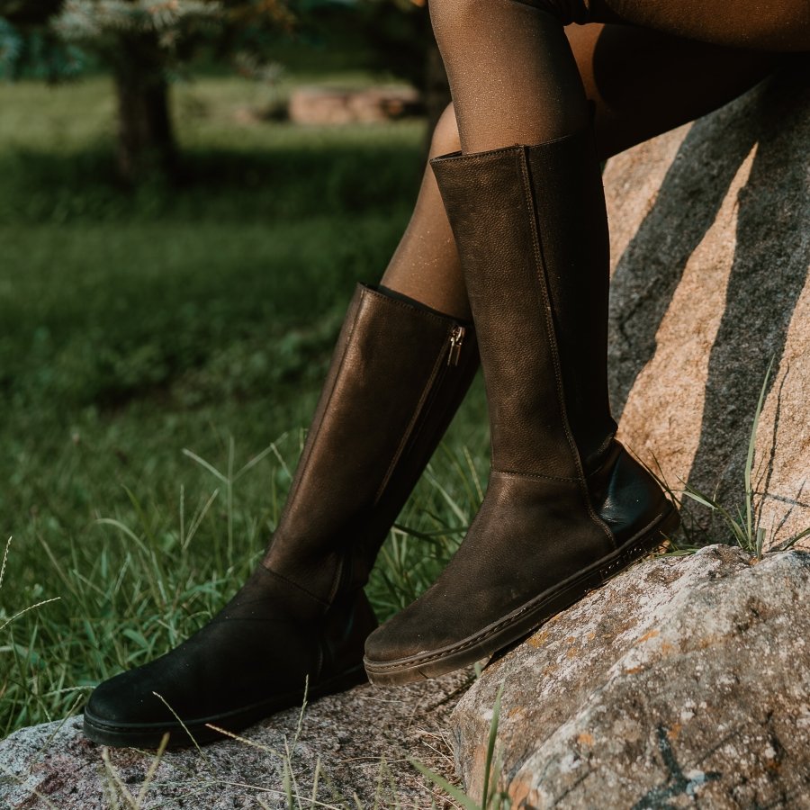 A close up of a pair of tall leather Peerko Regina barefoot riding style boots being worn by a person sitting on a large boulder.