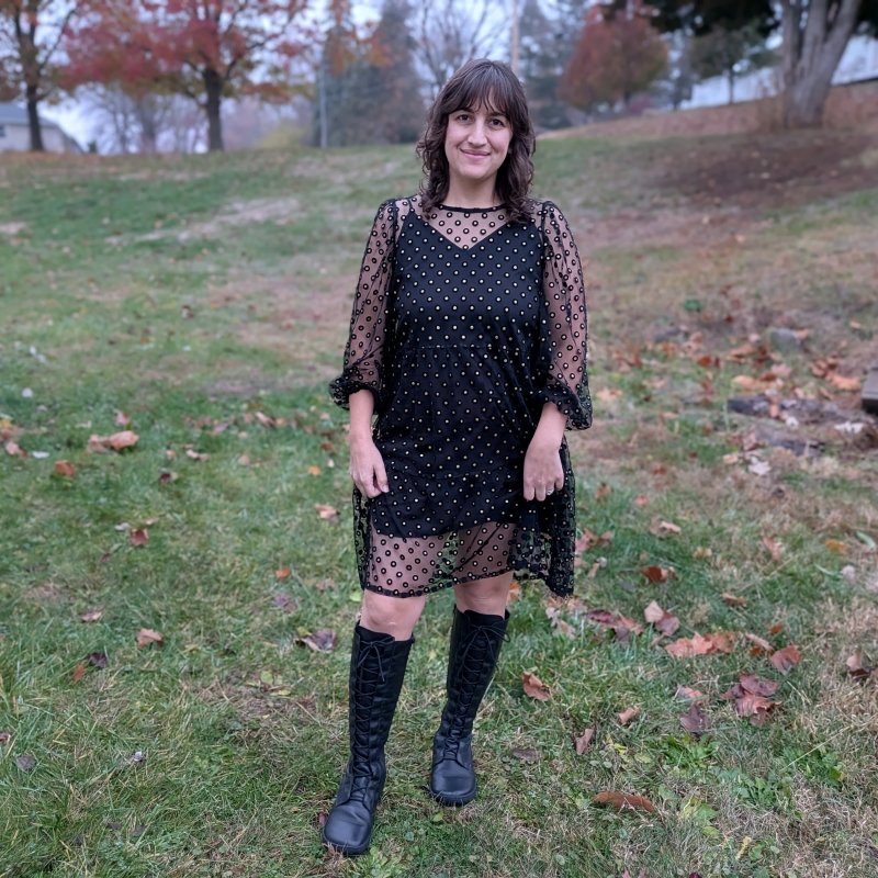 Full body view of a woman standing outside on a foggy day wearing a polka dot sheet overlay black dress and a pair of tall lace up leather barefoot boots from Shapen Barefoot. The Abby leather boot has a wide toe box and flexible outsole plus fleece lining.
