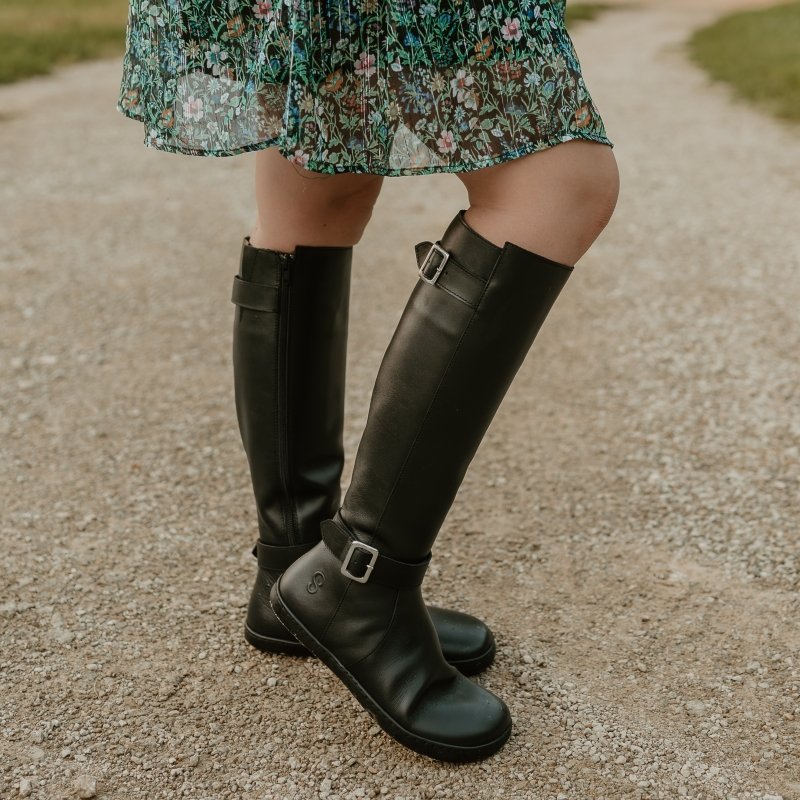 A side close up view of a pair of Shapen Glan tall black riding boots with buckle details and a flat flexible outsole. They are shown with a green floral dress peeking at the top of the frame.