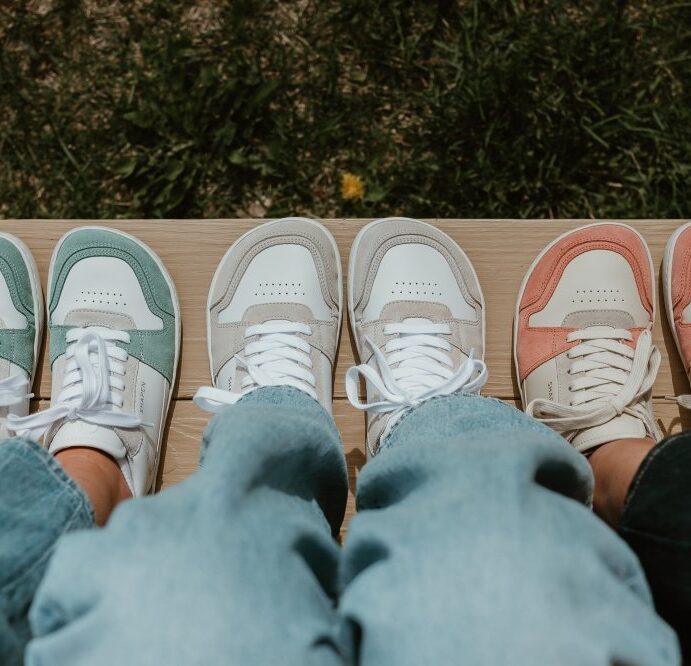 A line up of 3 people wearing Shapen ReWind soft leather barefoot sneakers in 3 different colors - Mint, gray, and peach. Their feet are resting on a bench and they are all wearing different denim washes