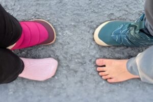 Top down view of 2 kids facing each other standing on ground covered in light snow. Each are wearing 1 snow boot on a foot, and the other foot is bare or socked to show the anatomical toe box of the barefoot Be Lenka and Vivobarefoot snow boots