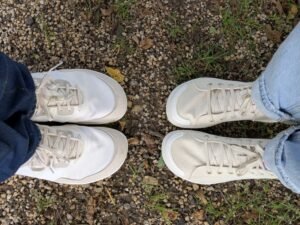 Two people's feet close up in Tolos wide toe box Transition models standing toe to toe on a grassy and gravel path.